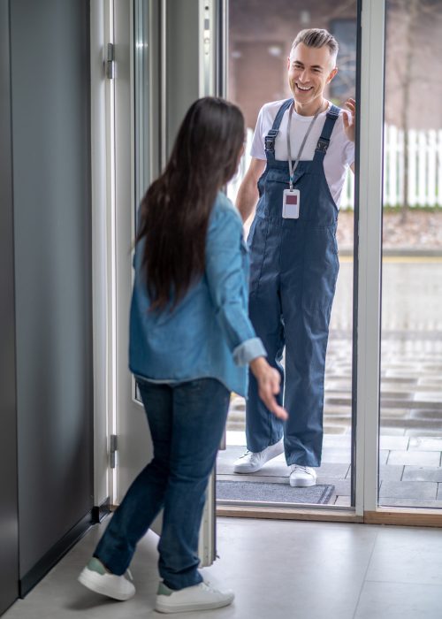 Invitation. Woman with long dark hair with her back to camera opening glass door inviting smiling man plumber in overalls into house